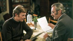 Two men sit at a dining table; one in a dark sweater leans in while the other in a suit reads a paper menu or document to him.