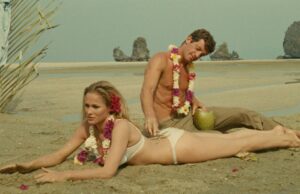 Man and woman in flower leis lounge on a sunny beach, with water and rock formations in the background.