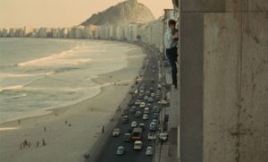 Man clinging to the side of a tall building overlooking a busy beachfront street, with cars on the road and high-rise apartments along the shore, and a rocky hill in the background.