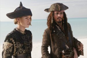 Two people dressed as pirates stand on a beach with the sea in the background, the woman in ornate black-gold clothing and a large hat, the man in a weathered coat with a tricorne hat.