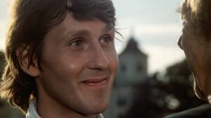 Close-up of a smiling young man looking at someone off-screen, outdoors with a blurred cityscape in the background.