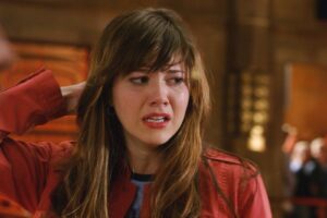 Woman with long brown hair in a red jacket crying, tears on her cheek, looking to the side in a dim indoor setting.