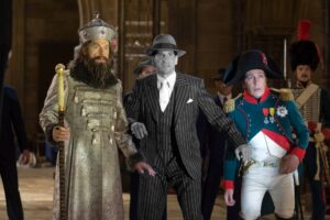 Three costumed men walk forward: a silver-robed king holding a jeweled staff on the left, a man in a pinstripe suit and fedora in the center, and a blue military uniformed officer on the right.
