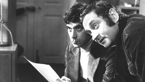 Two men lean in to read a sheet of paper, studying it closely in a black-and-white photo.