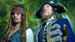 Two men in 18th‑century pirate attire gaze upward, one with a red bandana and braids, the other in a blue coat with gold embroidery, standing outdoors.