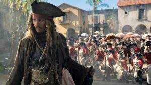 Pirate with a tricorn hat and dreadlocks stands apart as soldiers march in red coats behind him in a sunlit colonial town square