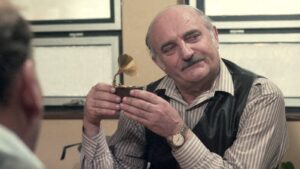 Older man with a mustache holding a small antique gramophone, smiling at someone off-camera in an office setting.