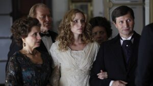 Bride in a white lace gown stands with two older guests in formal attire, looking off-camera.