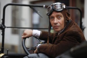 Man in vintage leather aviator helmet and goggles sits in an open-top car, looking at the camera with a calm expression.
