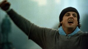 Man in a dark beanie and light blue scarf shouts with arms raised, facing the camera.