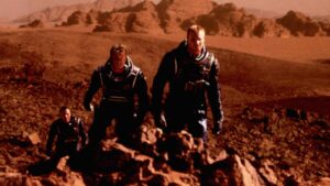 Three astronauts in bulky suits walk over rocky, reddish terrain with distant cliffs, resembling a Martian landscape.