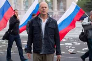 Central man with a serious expression in a dark jacket stands in a street surrounded by people waving large Russian flags at a protest.