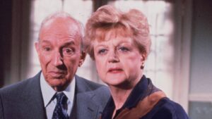 Two older adults, a man in a suit and a woman in a dark blazer, look intently at the camera.
