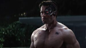 Muscular man shirtless with a damaged robotic cheek and a red glowing eye, outdoors at night.