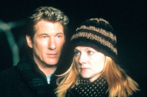 A man and woman pose close together at night, both looking off to the side, wearing dark jackets and a knitted hat for the woman.