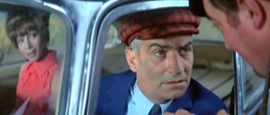 Man in a blue suit and red patterned cap leans toward a car window, talking to a man inside; a woman in pink sits in the backseat watching.