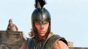 Male warrior wearing a bronze helmet with a plume and armor, staring forward on a battlefield.