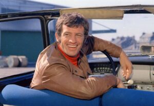 Smiling man in a brown jacket sits in a blue convertible, with the dashboard and steering wheel visible behind him.