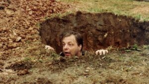 Man emerging from a muddy hole in the ground, reaching up with hands.