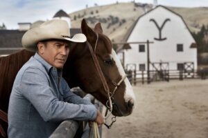 Man in a white cowboy hat leans on a railing with a brown-and-white horse at a ranch.”,