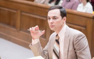 Man in a light brown plaid suit holds a pen while seated in a courtroom.
