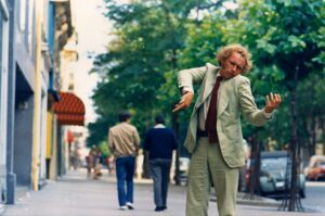Man in a light suit and red tie perform a dance on a city sidewalk while pedestrians pass in the background.
