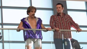Two men lean on a glass railing in a bright, modern atrium; the left man wears a metallic purple sleeveless jacket and silver shorts, striking a pose while the right man in a red plaid shirt looks on.