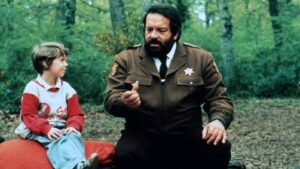 Bearded man in a brown sheriff uniform kneels in a forest, handing a small object to a young boy seated on a red cushion.