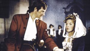 Pair in historical costumes share a moment: a man in a red coat and a woman in a bonnet and gown meet in a formal setting.