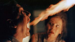 A man shouts with mouth wide open toward another man as a bright flame crosses the frame between them.